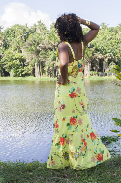 Nainen hallitseva vihreä värillinen mekko poseeraa valokuva puiden joukossa kasveja ja kukkia ja joki taustalla. Ilon, kypsyyden ja mustan tietoisuuden käsite. - Valokuva, kuva