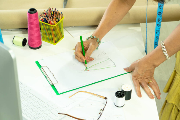 Aus nächster Nähe zeichnet eine reife Kostümbildnerin Skizzen auf Papier. Asiatische Senior-Schneiderin Inhaber eines kleinen Geschäfts, das am Arbeitsplatz in einem Modegeschäft arbeitet. Beruf Kreative Textilindustrie. - Foto, Bild