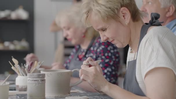 Eine Gruppe älterer Menschen bei einem Meisterkurs in Töpfern formte und schnitt gemeinsam eine Zeichnung auf Tassen aus Ton für die Herstellung von Keramikgeschirr - Filmmaterial, Video