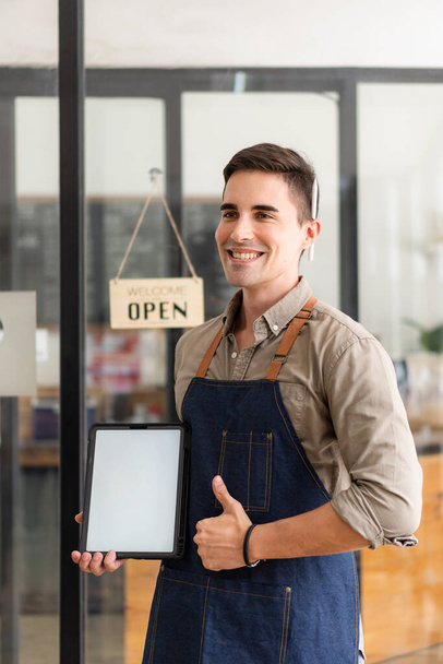 Ein gutaussehender junger Mann steht neben einem Schild vor der Ladentür und hält ein Tablet mit weißem Bildschirm in der Hand.. - Foto, Bild