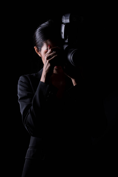 Азиатка фотограф держать камеру с внешней точкой вспышки для съемки темы, носить официальный костюм. студия освещения черный фон изолированных копий пространства, репортер журналист взять фото знаменитость - Фото, изображение
