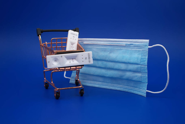 Covid-19 a koronavirus zdravotní a nákupní koncepce s maskou, nákupní košík a rychlé protilátky testovací sady přes modré pozadí - Fotografie, Obrázek