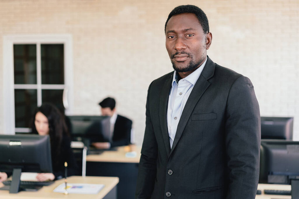 Αυτοπεποίθηση Αφροαμερικανός επιχειρηματίας στέκεται στο γραφείο με θολή υπαλλήλους που εργάζονται στο γραφείο τους στο παρασκήνιο. Πορτρέτο Αφροαμερικανού επιχειρηματία. - Φωτογραφία, εικόνα