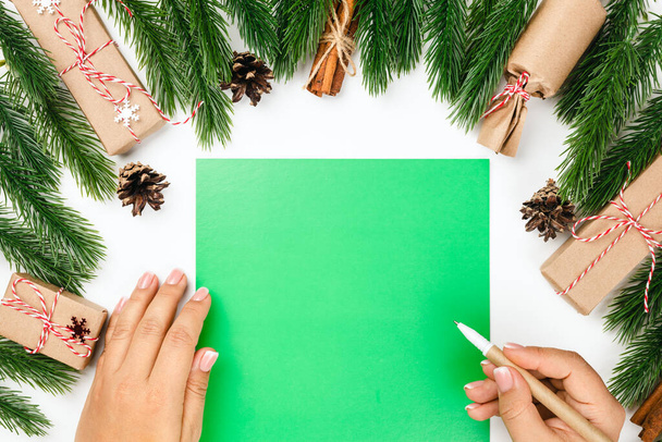 top view close-up female hands holding green cardboard with copy space card for New Year greetings in frame of fir branches and gift boxes - Fotografie, Obrázek