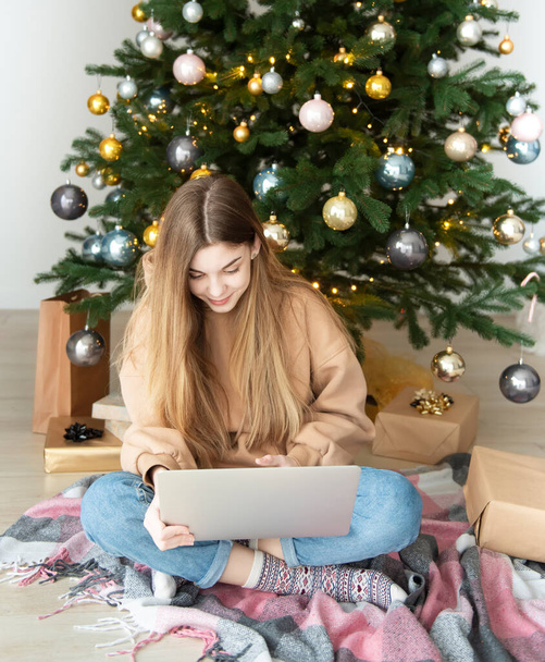 Tinédzser lány ajándékokkal és laptoppal a karácsonyfa mellett. Nappali belső tér karácsonyfával és dekorációval. Újév. Ajándékozás. - Fotó, kép