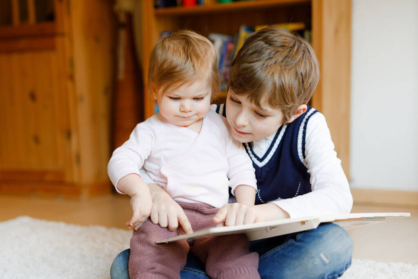 Un garçon de l'école lisant un livre pour une petite fille en bas âge, deux frères et sœurs assis ensemble et lisant des livres. Belle belle famille amoureuse, mignon bébé et enfant s'amuser à la maison, à l'intérieur. - Photo, image