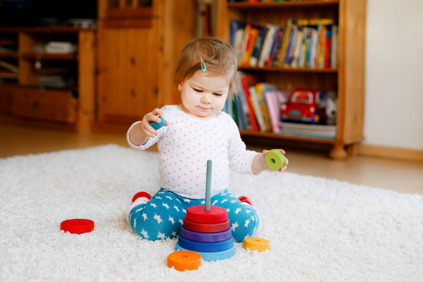 Адекватная симпатичная маленькая девочка, играющая с учебными деревянными игрушками дома или в детском саду. Туфелька с разноцветной стопкой пирамид и музыкальной игрушкой. Счастливый здоровый ребенок веселится с разными игрушками - Фото, изображение