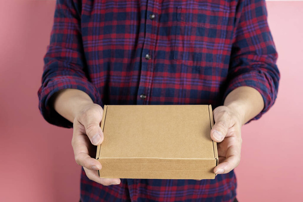 A cardboard box in the hand of a man wearing a dark red plaid shirt. A front view of the courier extending the parcel in front of him. delivery concept - Фото, изображение