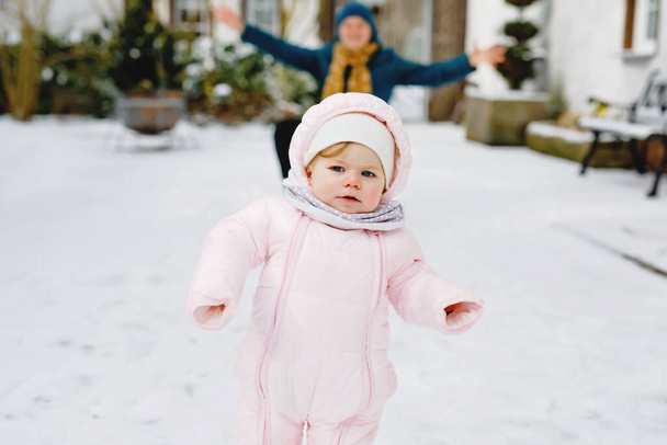 Lovely little baby girl making first steps outdoors in winter. Cute toddler learning walking. Happy mother on background. Beautiful family, mum and daughter on snowy day, walk through snow. - Photo, Image