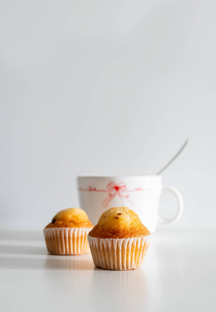 A vertical shot of a white mug and small chocolate chip muffins sprinkled with cocoa on white background with copy space - Foto, afbeelding