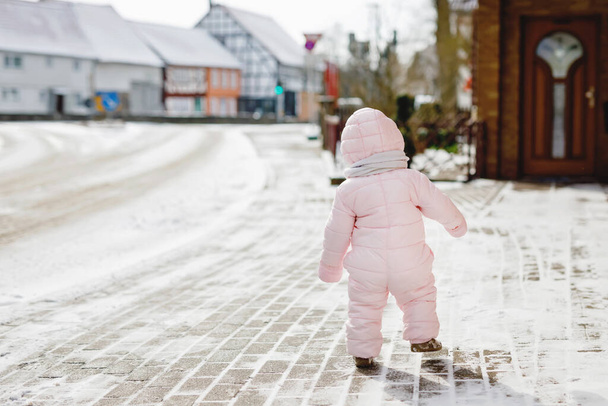 Boldog kislány teszi az első lépéseket a szabadban télen a hóban. Aranyos kisgyerek, aki járni tanul. A gyerek jól érzi magát a hideg havas napokon. Meleg baba rózsaszín ruhát visel hóvédőt és bólogatós kalapot.. - Fotó, kép