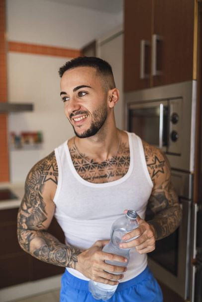 A vertical shot of a tattooed Caucasian male athlete drinking water after a workout at home - Foto, afbeelding