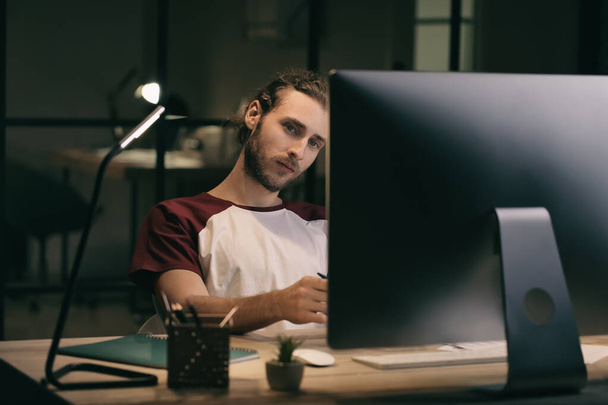 Молодой человек за компьютером в офисе поздно вечером - Фото, изображение