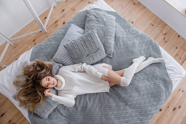 A millennial young woman with curly blond hair in white knitted sweater dress, leggings lies on large sofa with gray plaid, pillows. Christmas cozy morning in a stylish interior. Top view, copy space - Photo, Image