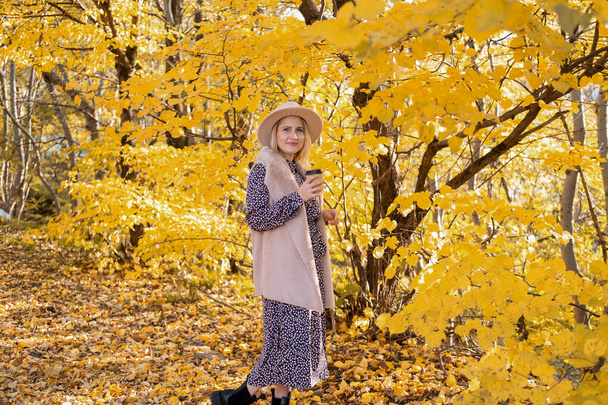 belle femme en vêtements à la mode et chapeau boit du café dans la nature à l'automne - Photo, image