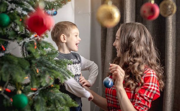 Mom with her child is decorating together the Christmas tree with toys and balls. Concept of family preparation for the New Year, cozy atmosphere and festive mood. - Photo, Image