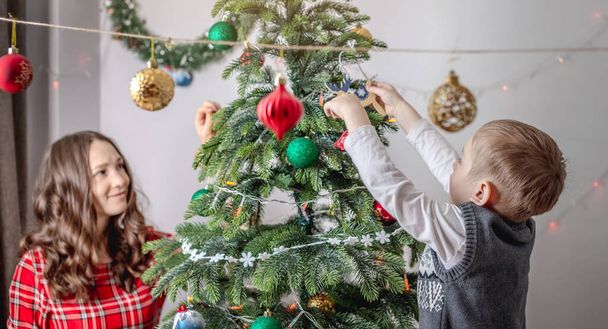 Mom with her child is decorating together the Christmas tree with toys and balls. Concept of family preparation for the New Year, cozy atmosphere and festive mood. - Photo, image