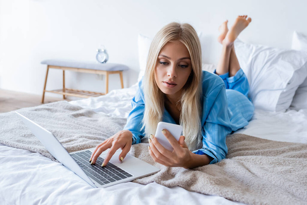 pleased young woman in pajamas using laptop and looking at smartphone in bedroom  - Foto, imagen