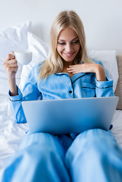 радостная молодая женщина в пижаме смотрит на ноутбук и держит чашку кофе  - Фото, изображение