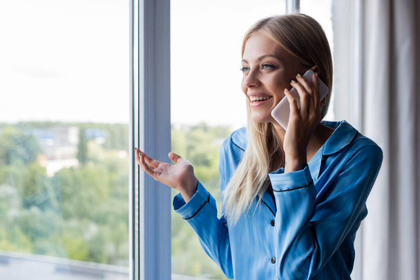 веселая молодая женщина разговаривает по сотовому телефону у окна  - Фото, изображение