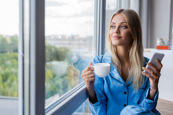 happy young woman holding cellphone and cup of coffee near window  - 写真・画像