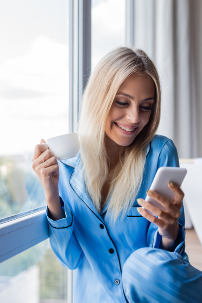 onnellinen nuori nainen tilalla älypuhelin ja kuppi kahvia ikkunan lähellä  - Valokuva, kuva