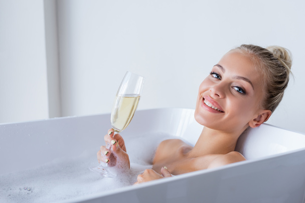young and joyful woman taking bath and holding glass of champagne  - Foto, Imagem