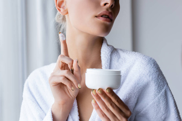 cropped view of young woman in bathrobe holding container and cosmetic cream on finger - Fotó, kép