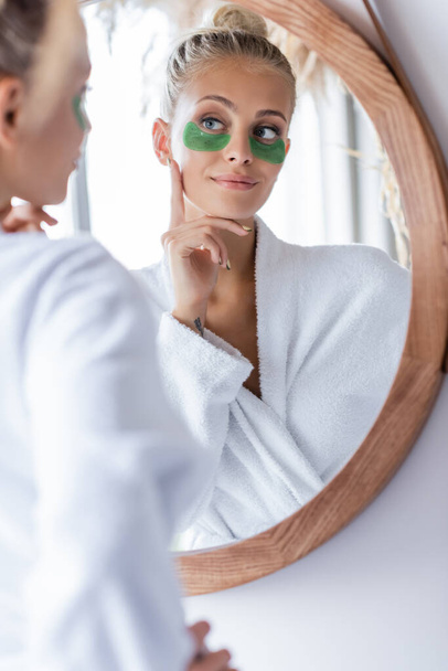 pleased young woman in bathrobe applying green eye patches near mirror  - Фото, изображение