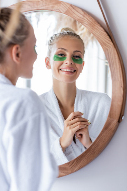 happy young woman in bathrobe with green eye patches smiling near mirror  - Foto, afbeelding