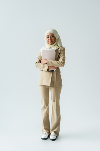 volledige lengte van gelukkig moslim vrouw in hijab en beige pak met mappen op grijs  - Foto, afbeelding