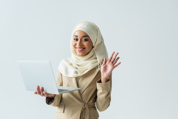 mulher muçulmana alegre em hijab e terno bege segurando laptop e mão acenando isolado em cinza  - Foto, Imagem