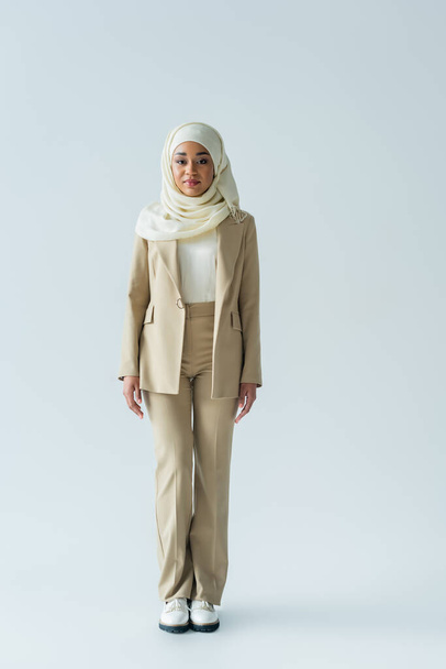 full length of muslim woman in hijab and beige suit standing on grey  - Φωτογραφία, εικόνα