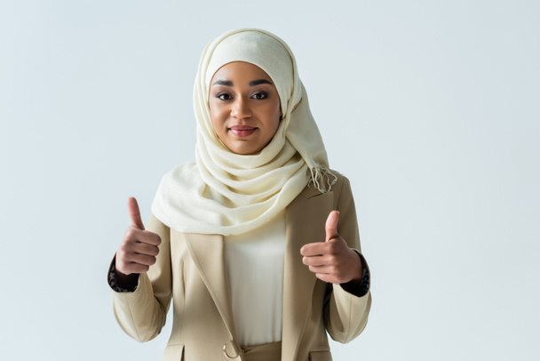 happy muslim woman in hijab showing thumbs up and smiling isolated on grey - 写真・画像