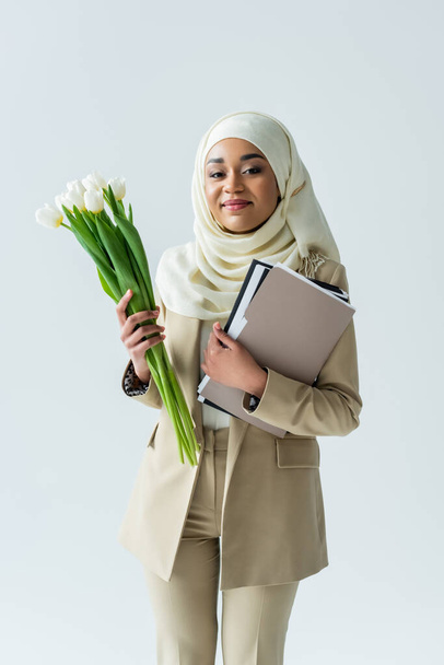 feliz musulmana mujer de negocios en hijab sosteniendo tulipanes y carpetas aislados en gris - Foto, Imagen