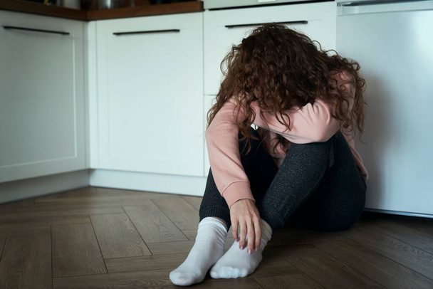 Broken young caucasian woman  with hidden face sitting on floor in the kitchen - Photo, Image