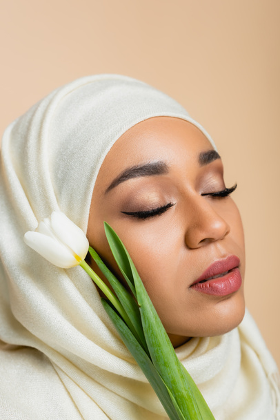 Portrait of pretty muslim woman in hijab with closed eyes near tulip isolated on beige  - Fotografie, Obrázek