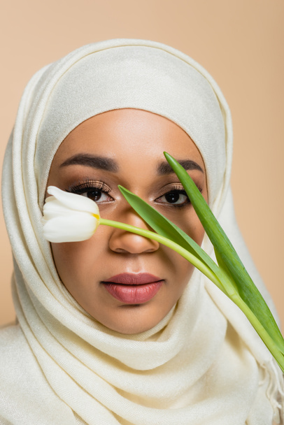 Portrait de jolie femme musulmane en hijab regardant la caméra à travers la tulipe isolée sur beige - Photo, image