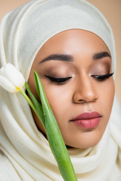 Ritratto di giovane donna musulmana in hijab vicino a tulipano isolato su beige  - Foto, immagini