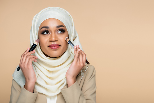 smiling muslim woman in hijab applying face foundation with cosmetic brushes isolated on beige - Foto, Imagen
