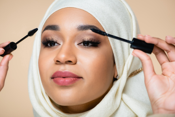 primer plano de la mujer musulmana en hijab aplicando rimel con cepillos aislados en beige - Foto, imagen