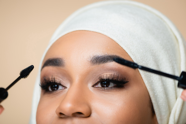 cropped view of muslim woman in hijab applying mascara with brushes isolated on beige - Foto, Imagen