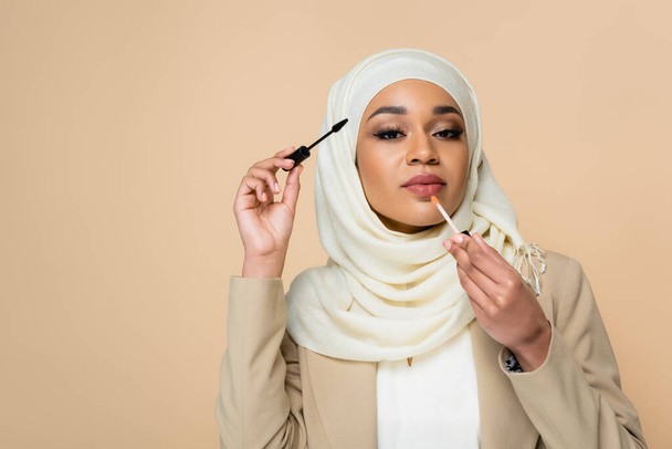 young muslim woman in hijab applying lip gloss and holding mascara brush isolated on beige - Foto, Imagem