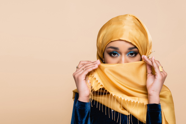 mujer musulmana con maquillaje de ojos brillantes cubriendo la cara con hijab aislado en beige - Foto, imagen