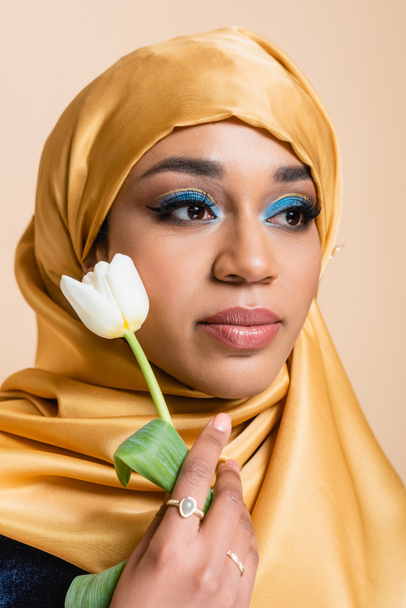 νεαρή μουσουλμάνα γυναίκα σε μαντίλα με φωτεινό μακιγιάζ κρατώντας τουλίπα απομονώνονται σε μπεζ - Φωτογραφία, εικόνα