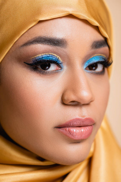 close up de jovem mulher muçulmana no hijab com maquiagem brilhante olhando para a câmera isolada no bege - Foto, Imagem