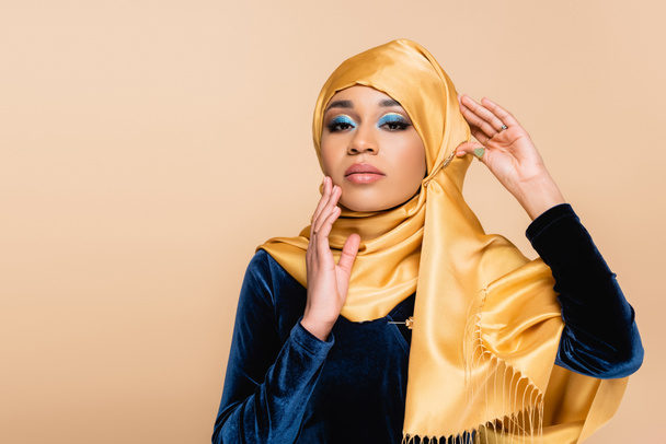 young muslim woman in yellow hijab with makeup looking at camera while posing isolated on beige - Foto, Bild