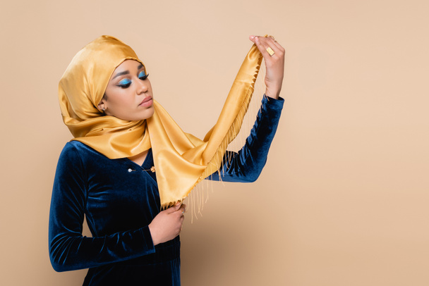 muslim woman with bright makeup holding headscarf isolated on beige - Foto, imagen