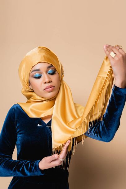 νεαρή μουσουλμάνα γυναίκα με φωτεινό μακιγιάζ κρατώντας μαντίλα απομονωμένη σε μπεζ - Φωτογραφία, εικόνα