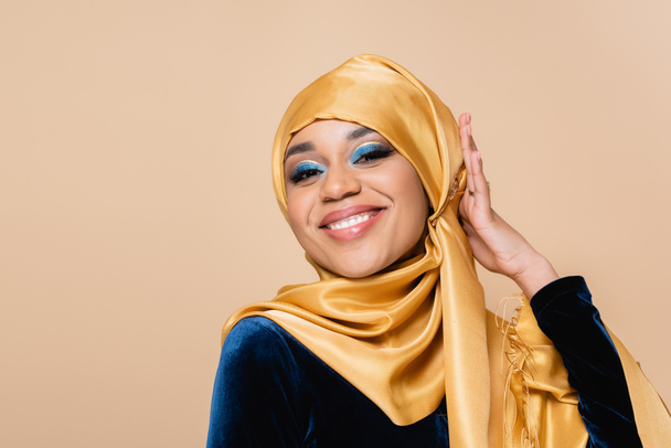 cheerful muslim woman adjusting yellow hijab isolated on beige - Foto, afbeelding